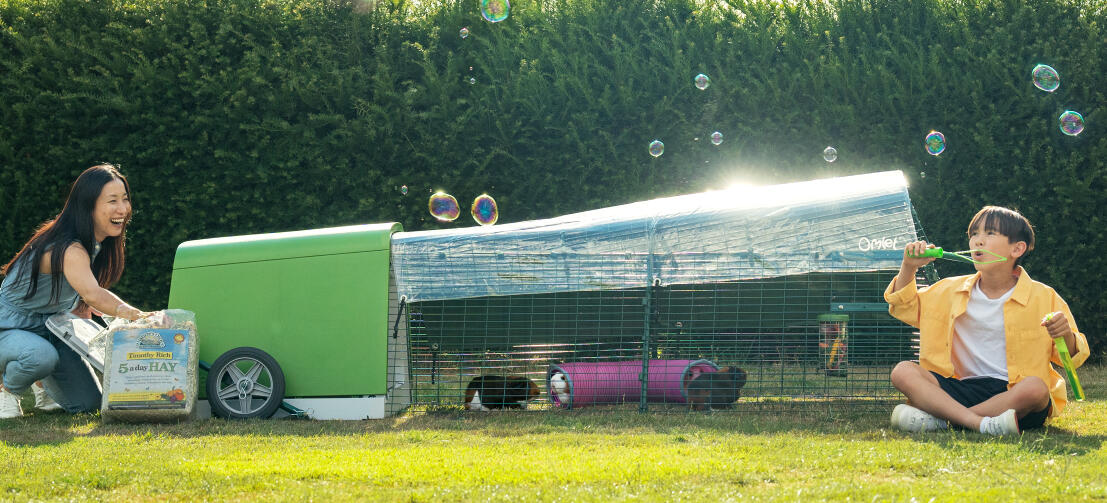 A child blowing bubbles in garden, while his mum is changing hay inside Eglu Go hutch.